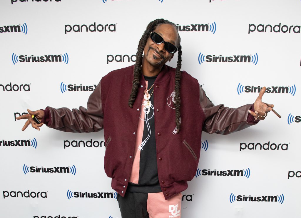 Snoop Dogg haussant les épaules et souriant devant un fond blanc, tenant un émoussé et un briquet