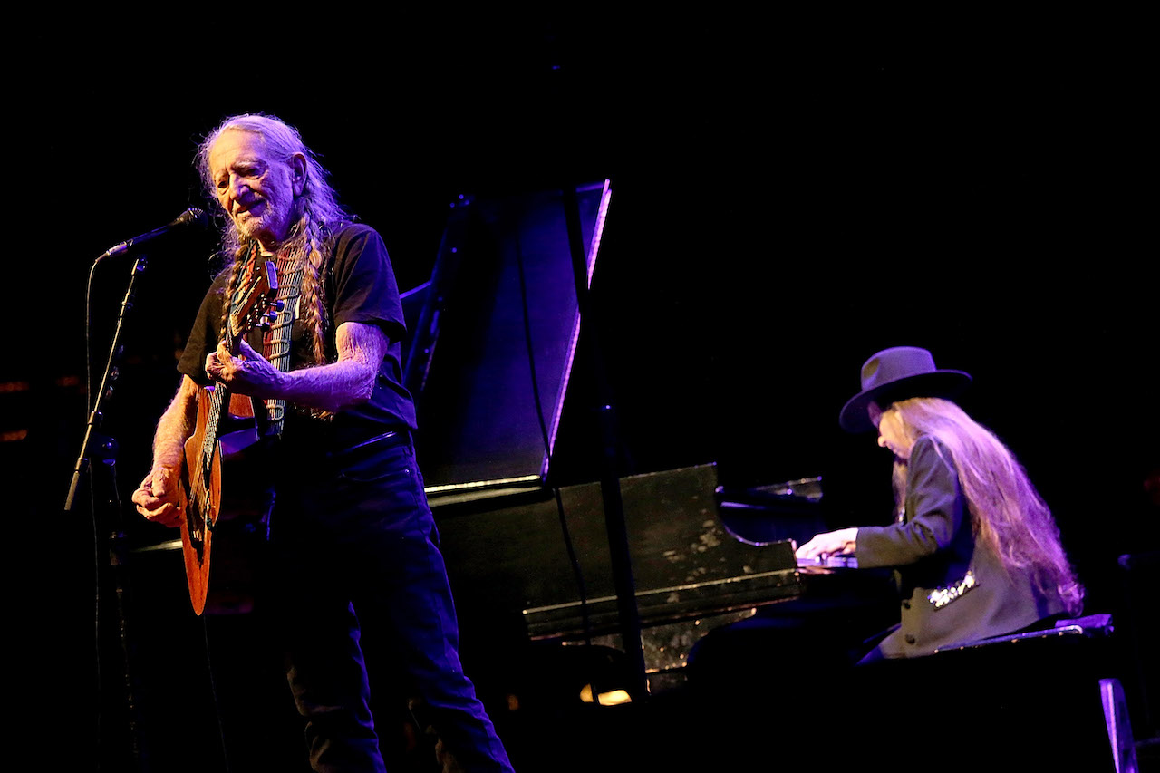 Willie Nelson se tient devant un microphone, tenant une guitare et Bobbie Nelson est assis au piano derrière lui