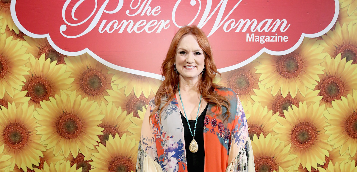 La femme pionnière Ree Drummond sourit vêtue d'une chemise noire et d'un pull multicolore devant une enseigne du magazine Pioneer Woman