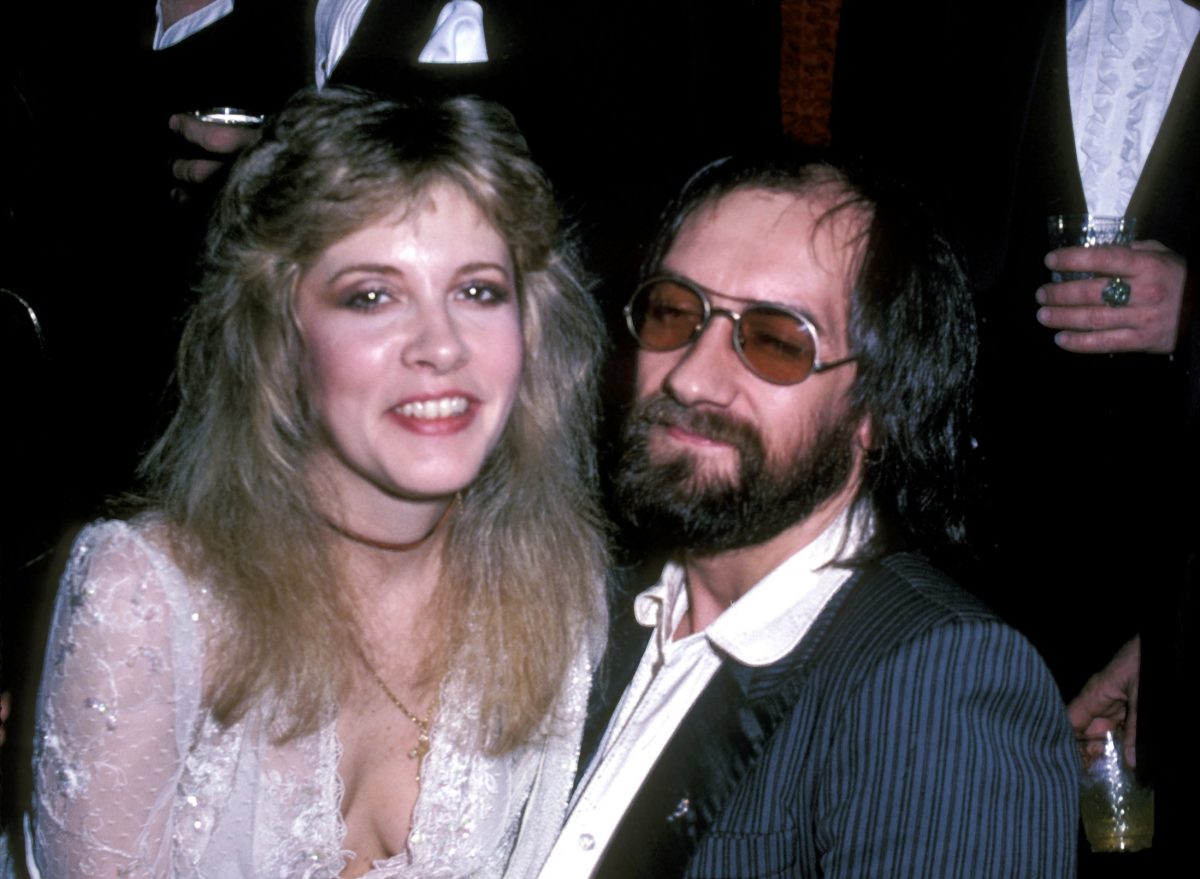 Stevie Nicks porte une robe blanche et est assise sur les genoux de Mick Fleetwood. Il porte des lunettes de soleil.