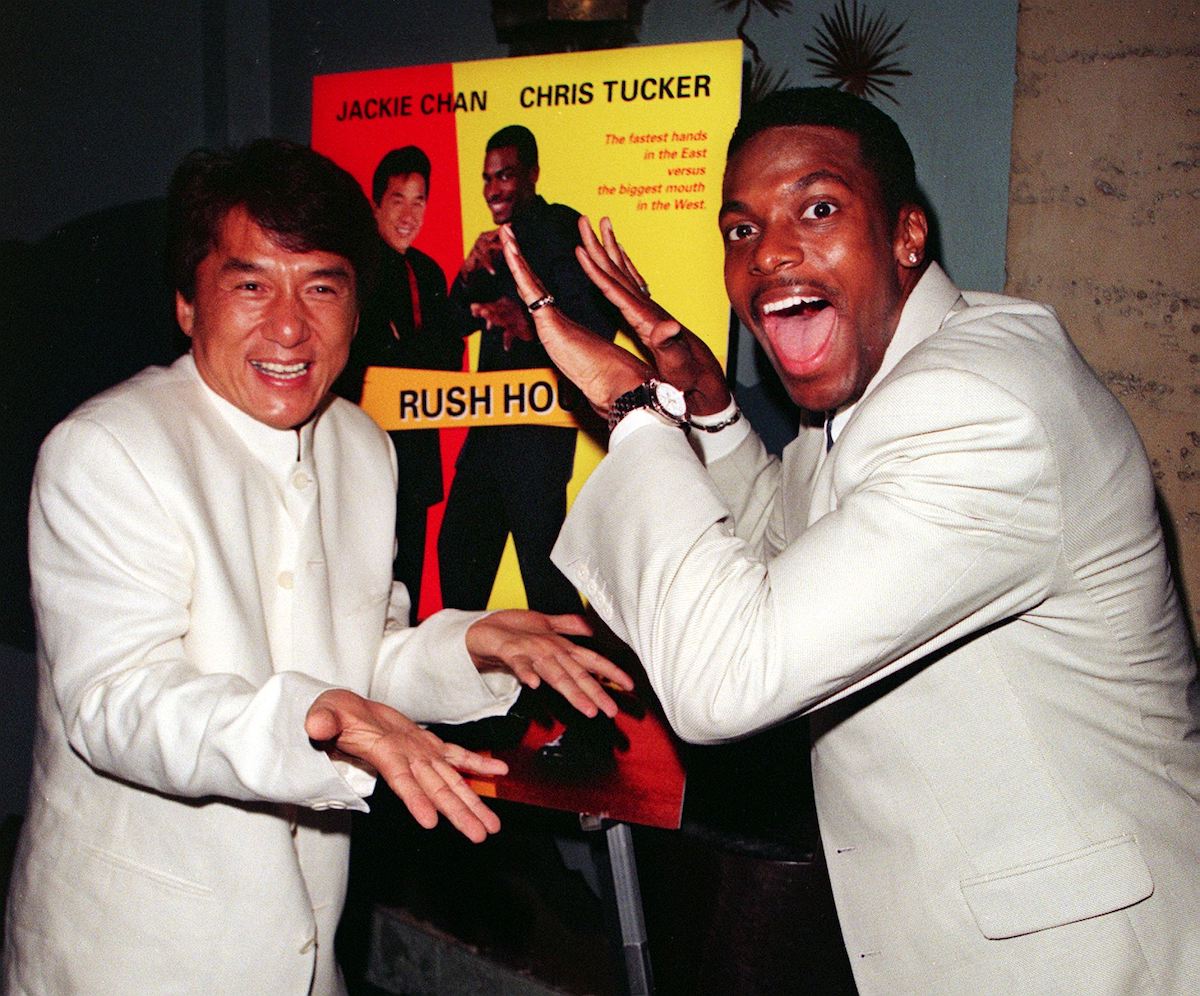 Jackie Chan et Chris Tucker souriants portent du blanc et un high five devant l'affiche 'Rush Hour'