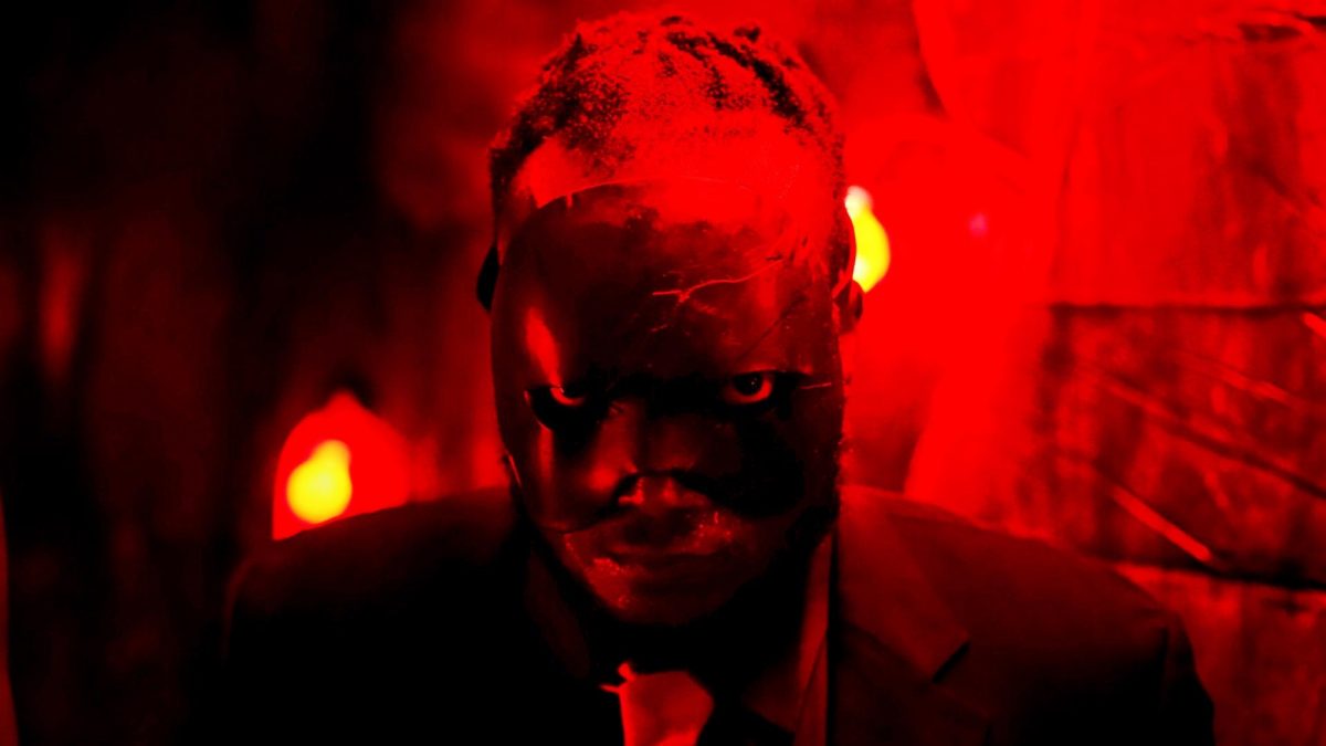 'Bitch Ass' Tunde Laleye dans le rôle de Bitch Ass portant un masque noir regardant la caméra
