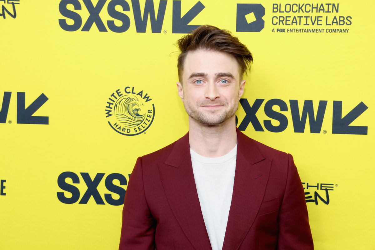 Daniel Radcliffe sur le tapis rouge à SXSW. Il se tient devant un mur jaune vif et porte une veste rouge et une chemise blanche.