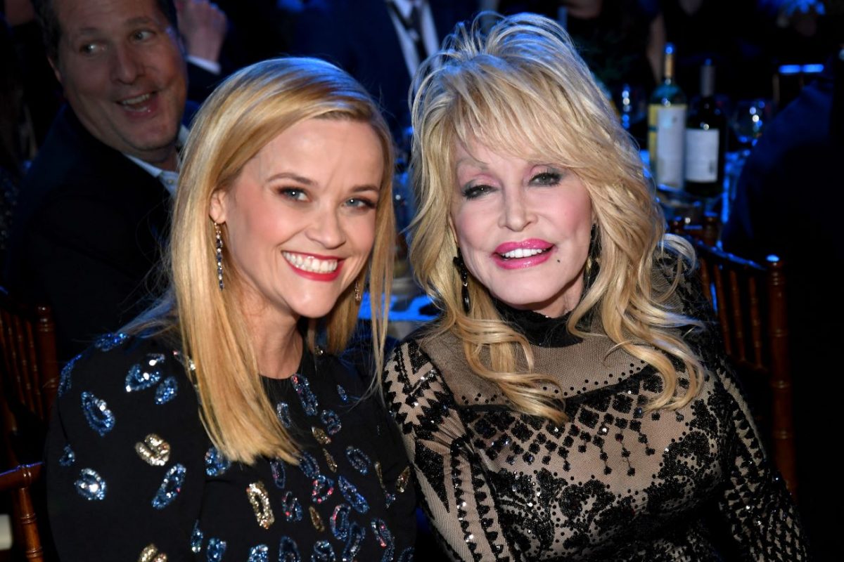 Reese Witherspoon et Dolly Parton sont assises l'une à côté de l'autre et sourient dans une pièce sombre.