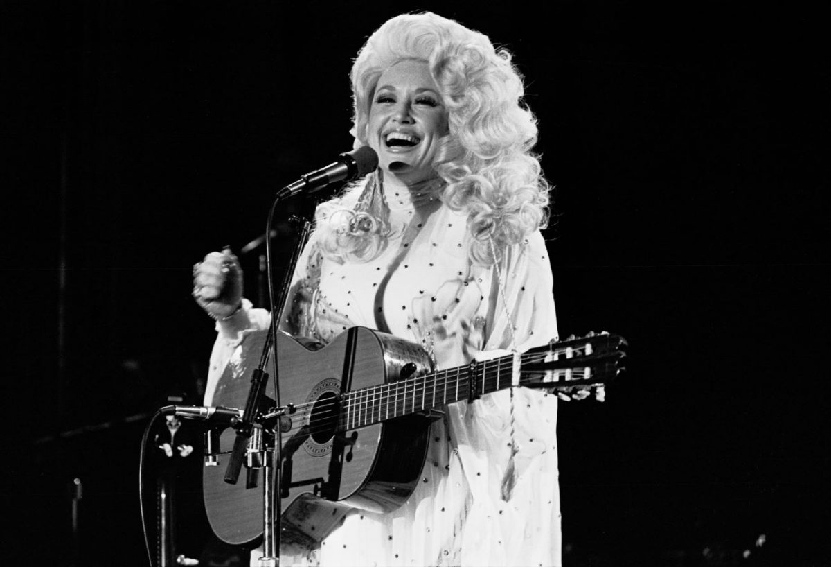 Une photo en noir et blanc de Dolly Parton tenant une guitare et debout devant un microphone dans les années 1970.