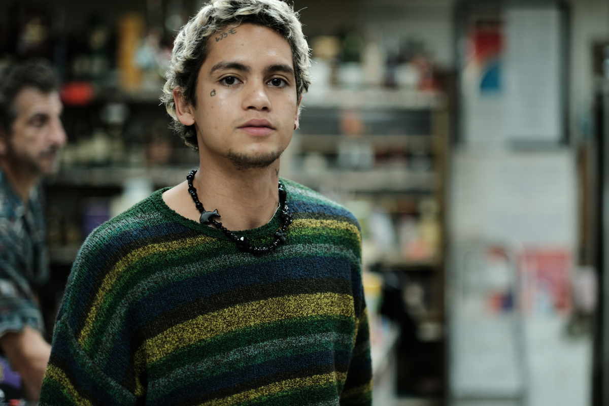 Dominic Fike dans le rôle d'Elliot dans Euphoria Saison 2. Elliot porte un collier et un pull rayé et se tient dans un dépanneur.