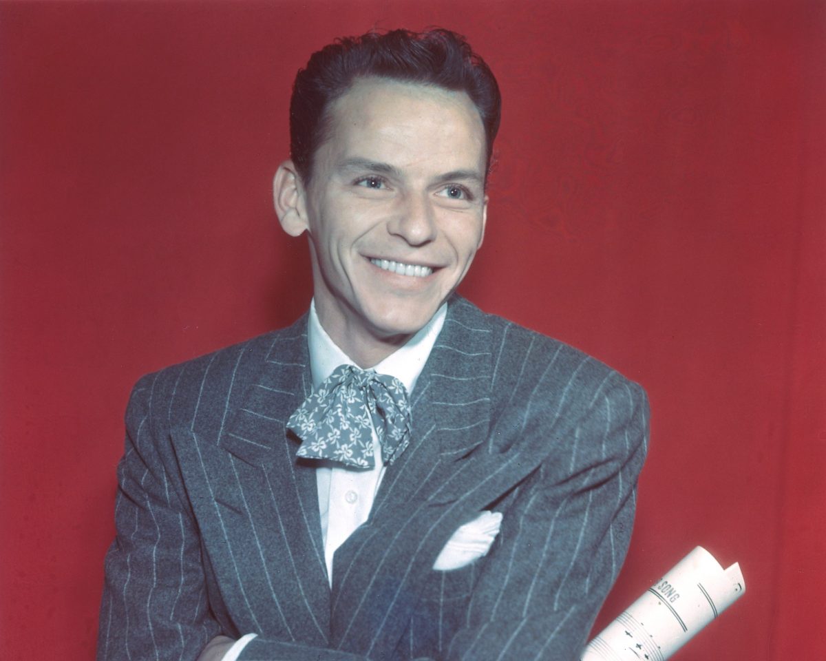 Frank Sinatra porte un costume et est assis devant un fond rouge. 