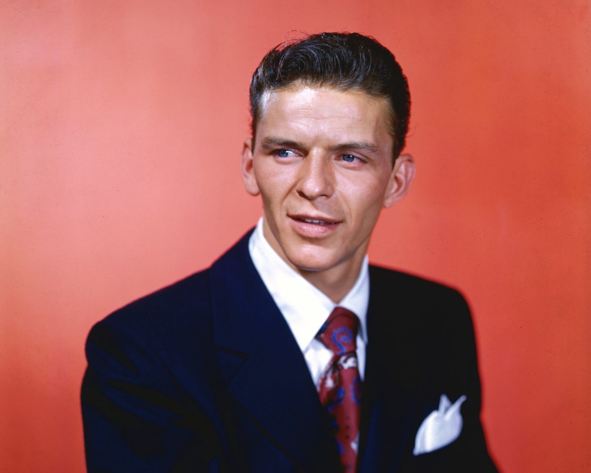 Frank Sinatra porte un costume et est assis devant un fond rouge.