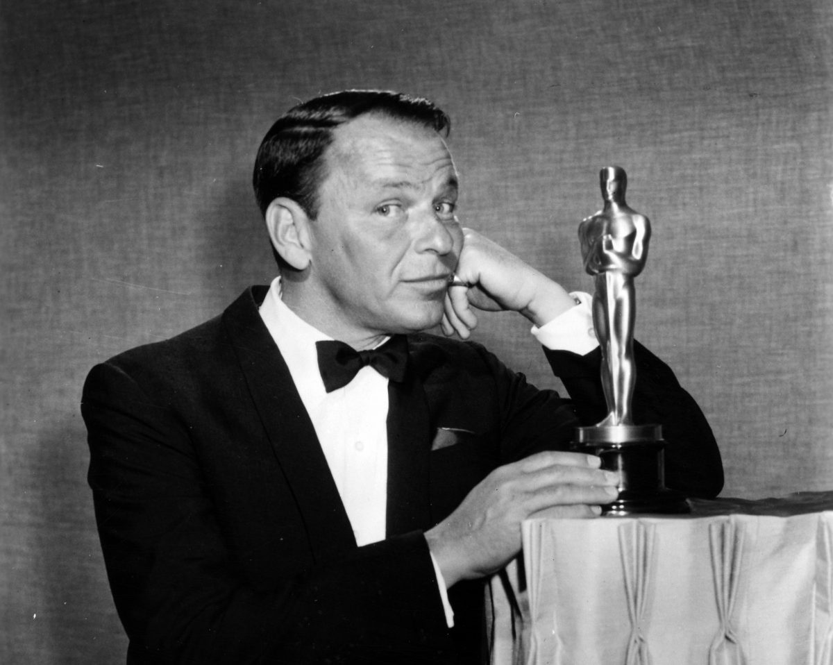 Une photo en noir et blanc de Frank Sinatra en smoking posant à côté d'une statue des Oscars.