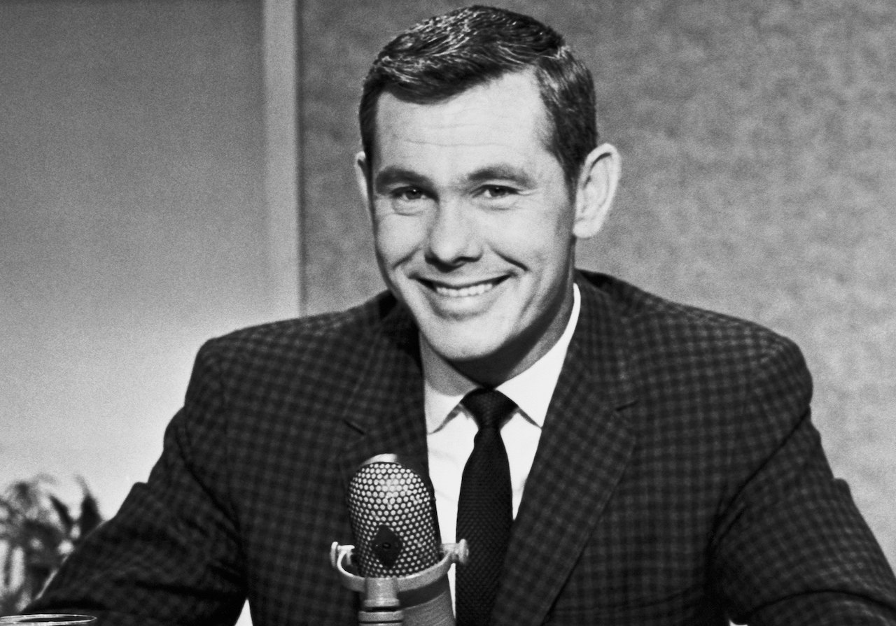 Photo en noir et blanc de Johnny Carson en costume, assis devant un micro et souriant c.  1963