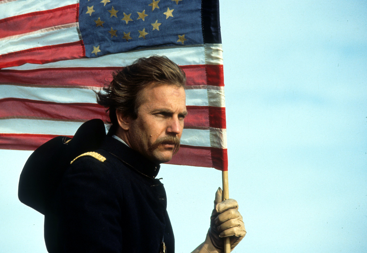 Kevin Costner tient un drapeau américain et regarde au loin