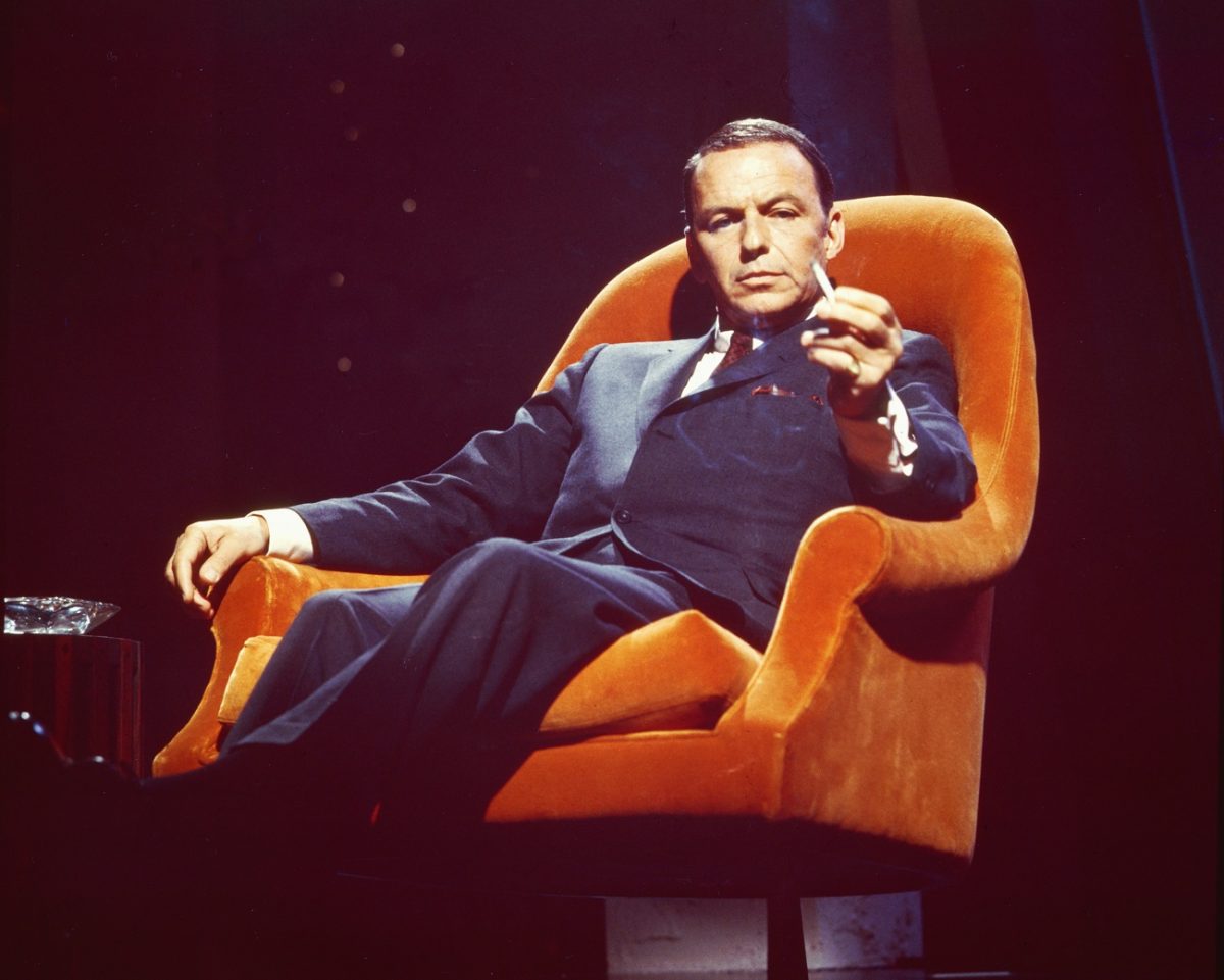 Frank Sinatra porte un costume et est assis dans un fauteuil orange.  Il tient une cigarette.