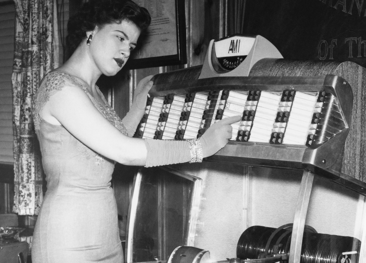 Photo en noir et blanc de Patsy Cline dans une blouse et des gants assortis, pointant vers un juke-box