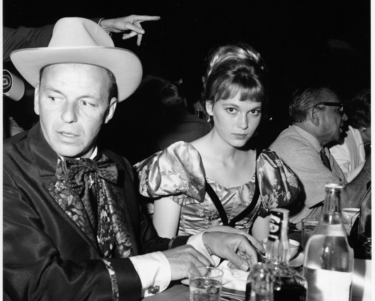 Une photo en noir et blanc de Frank Sinatra et Mia Farrow assis à une table.  Il porte un chapeau et elle porte une robe à manches bouffantes.