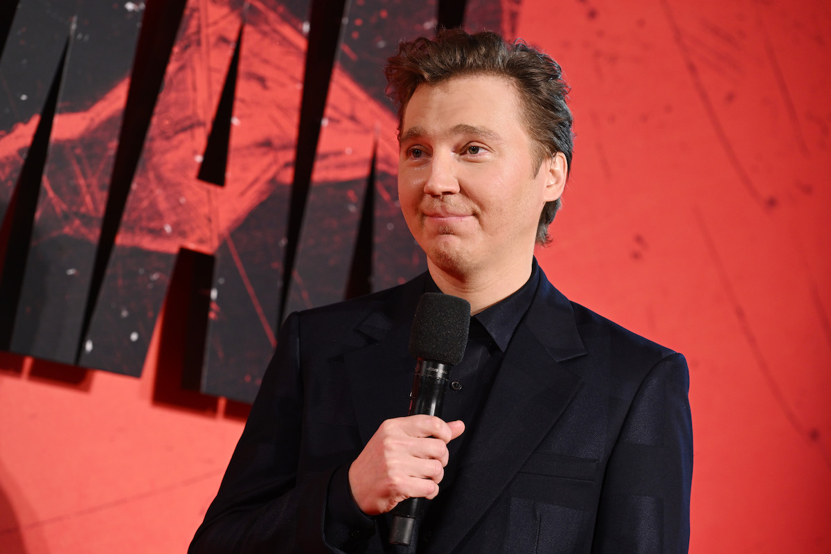 Paul Dano porte un costume noir et tient un micro sur le tapis rouge
