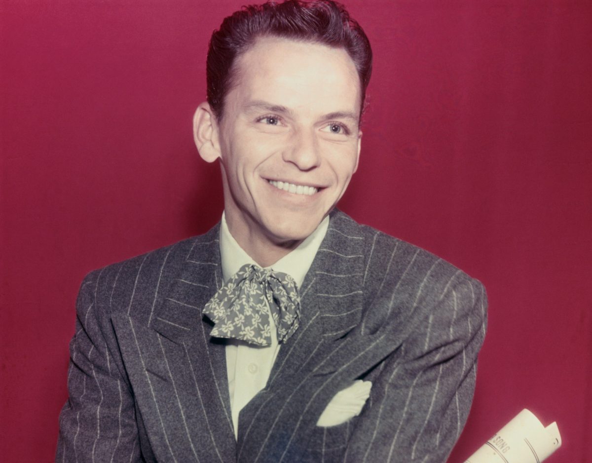 Frank Sinatra porte un costume gris et tient un papier enroulé. Il est assis sur un fond marron.