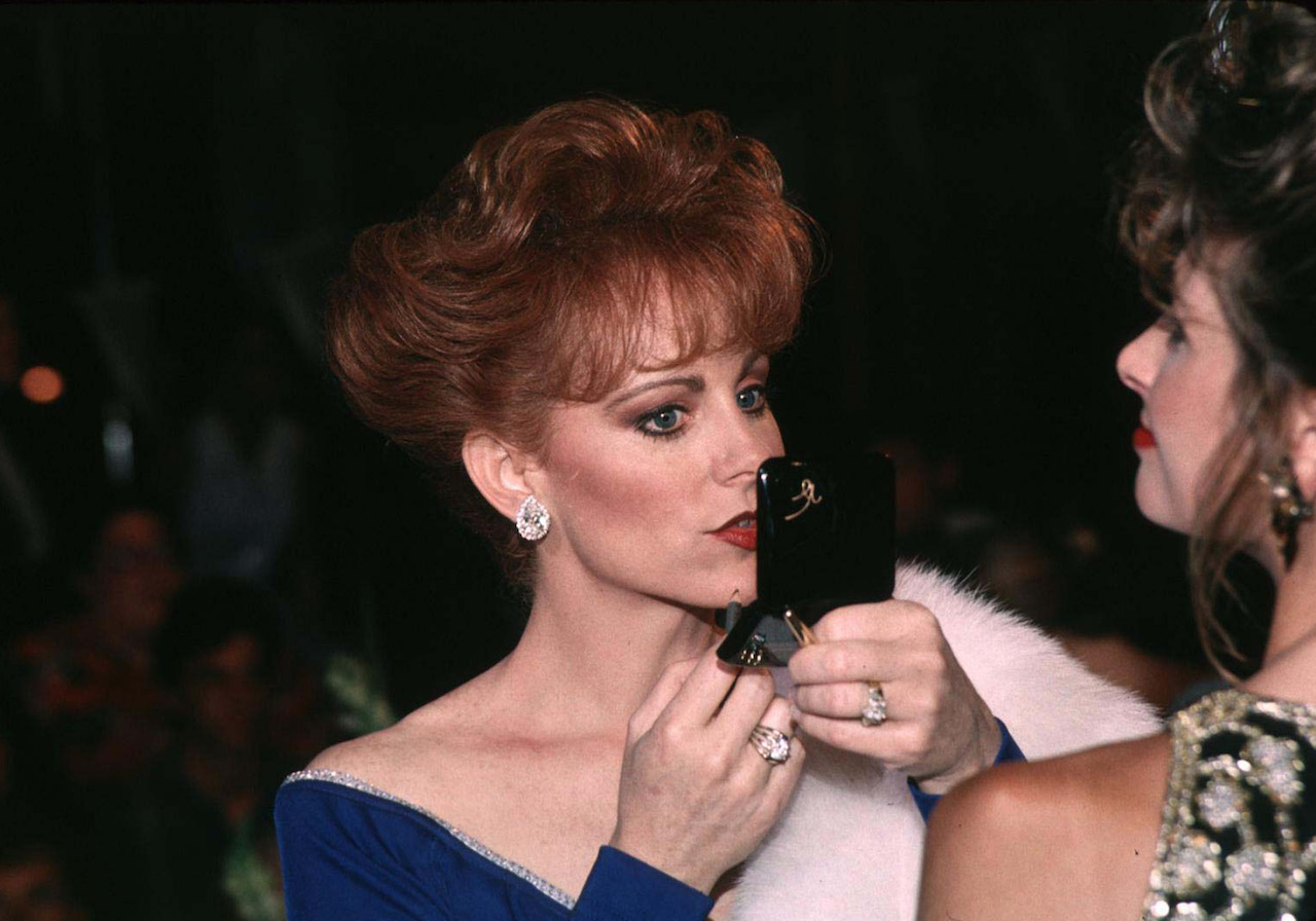 Reba McEntire dans une robe à épaules dénudées, se regardant dans un miroir et mettant du rouge à lèvres