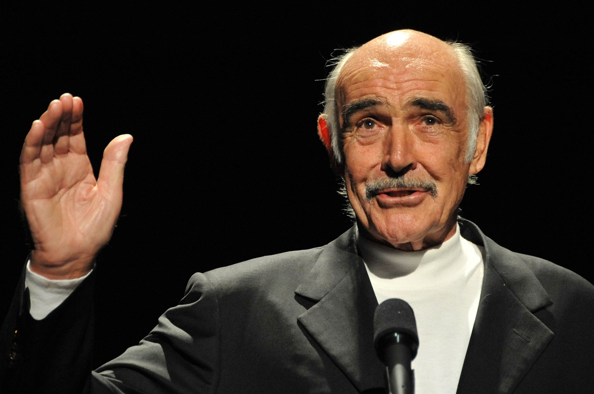 Sean Connery agitant tout en portant une chemise blanche à col roulé et un blazer.