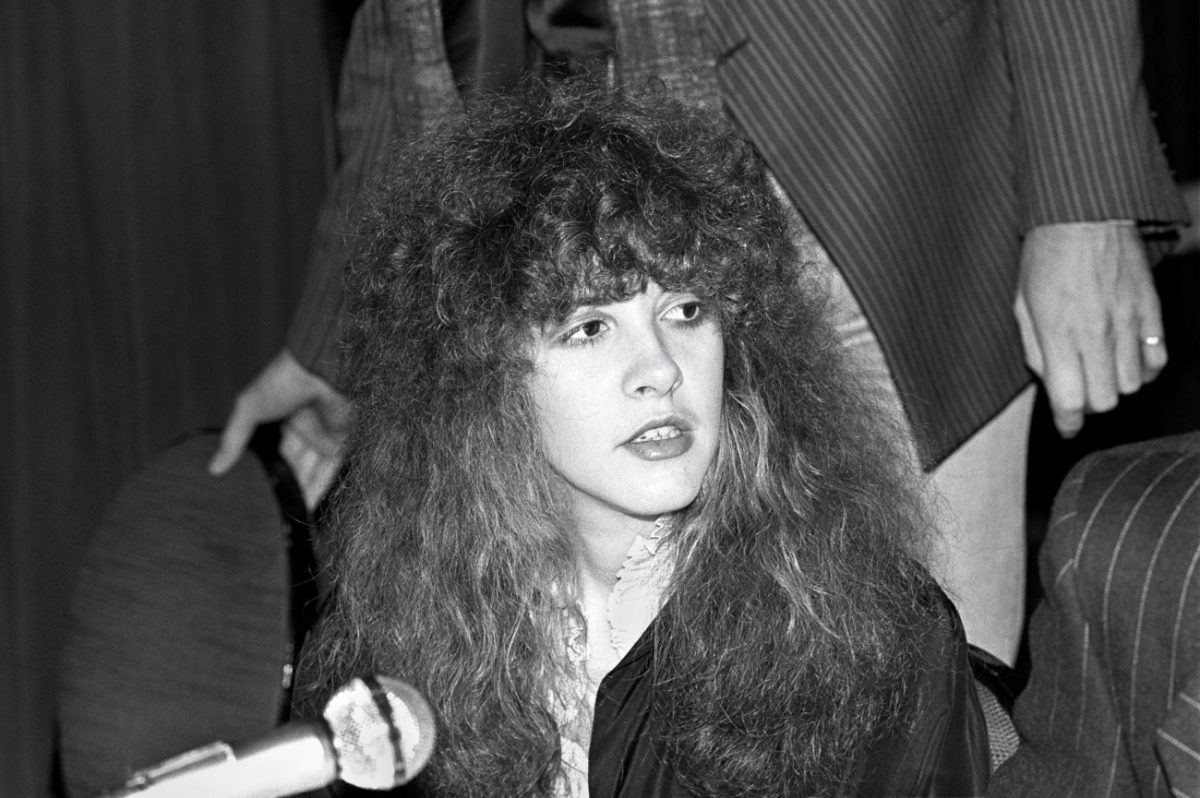 Une photo en noir et blanc de Stevie Nicks portant une chemise blanche et un pull noir et assis devant un microphone.
