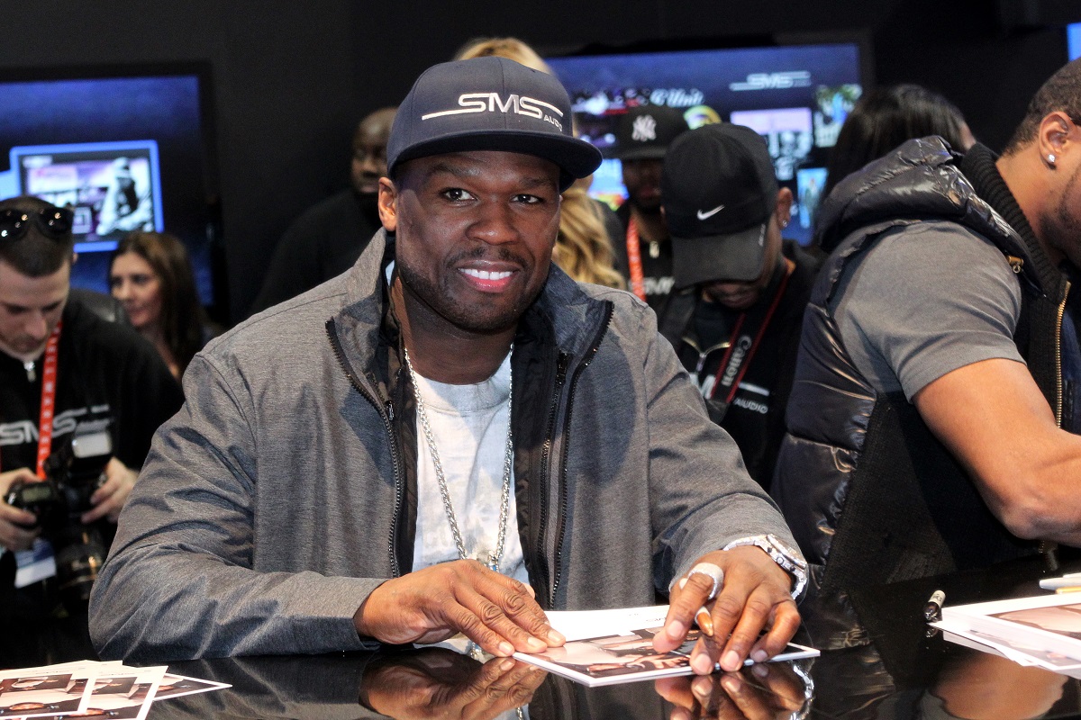 50 Cent sourit en portant une veste grise et une casquette.