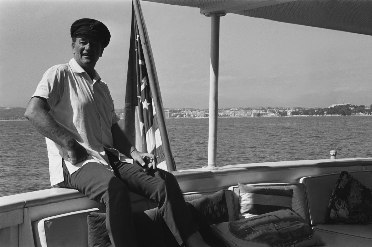 John Wayne à bord de Wild Goose, sa péniche, portant une chemise à col, un chapeau de capitaine, assis à côté du drapeau américain