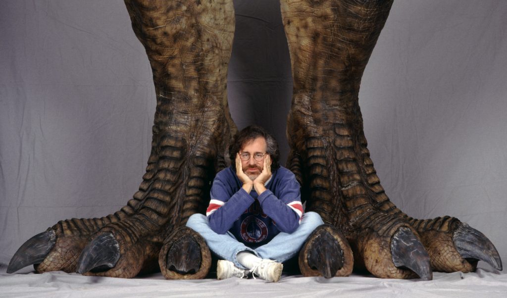 Steven Spielberg est assis entre une paire de pattes de dinosaures dans une séance photo faisant la promotion de «Jurassic Park»