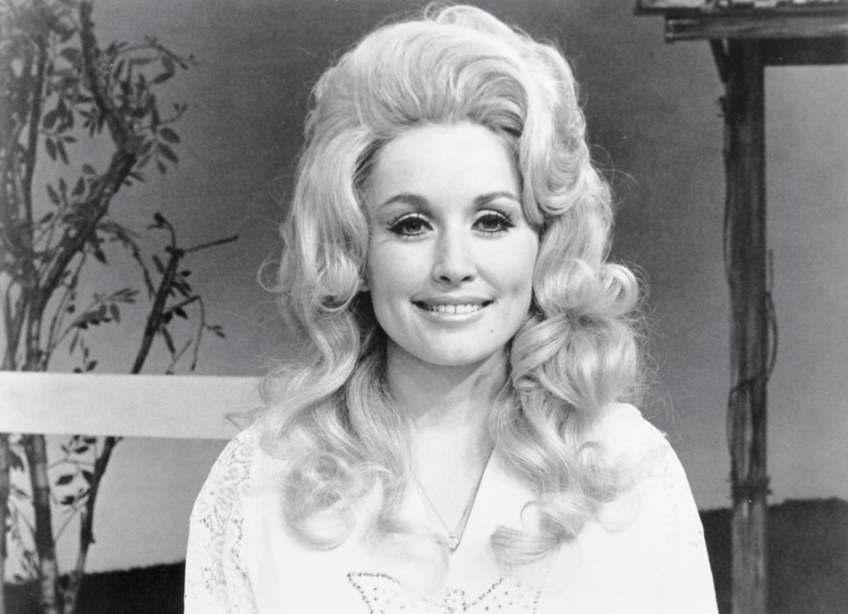 Une photo en noir et blanc de Dolly Parton assise devant une clôture et un arbre.