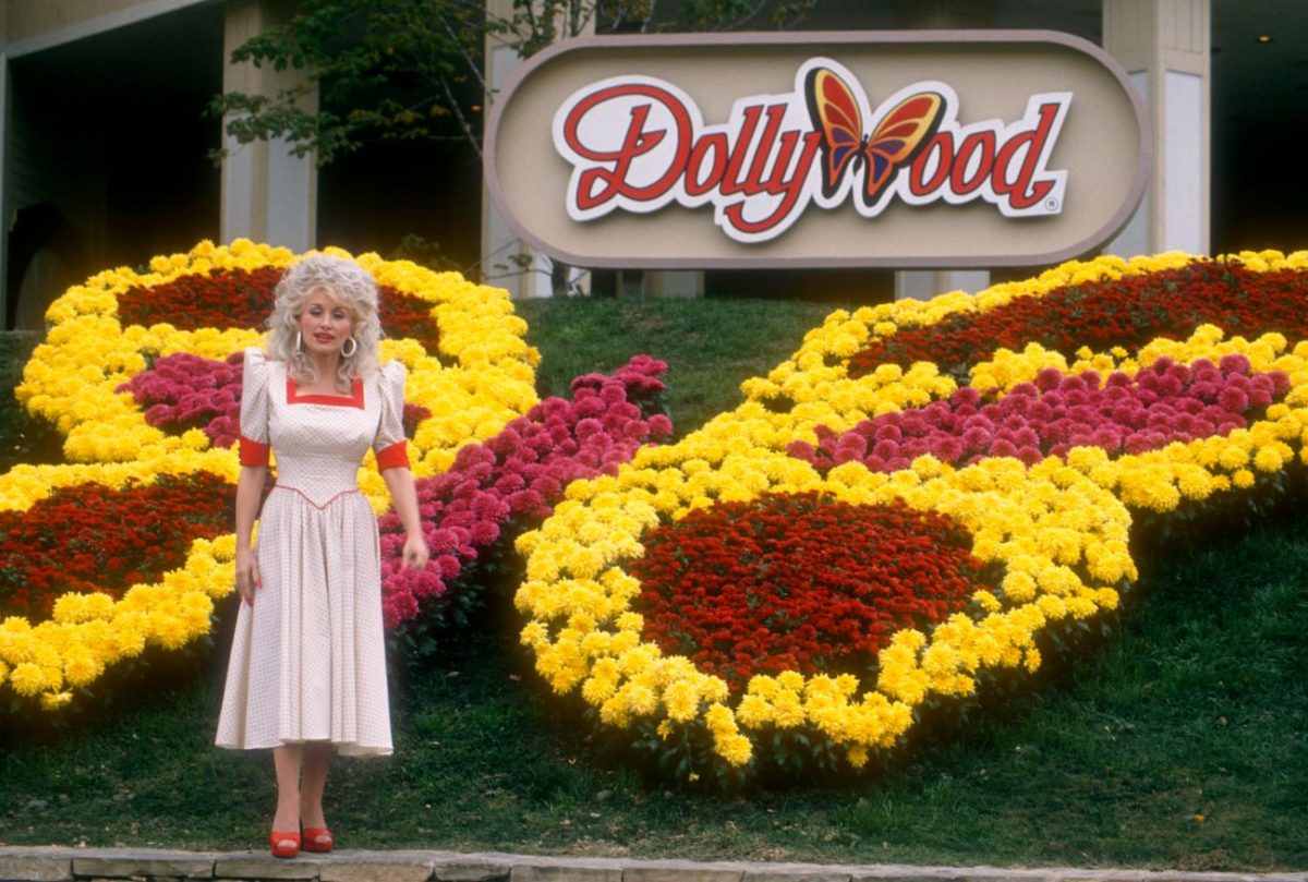 Dolly Parton se tient devant le panneau Dollywood et des fleurs plantées en forme de papillon.