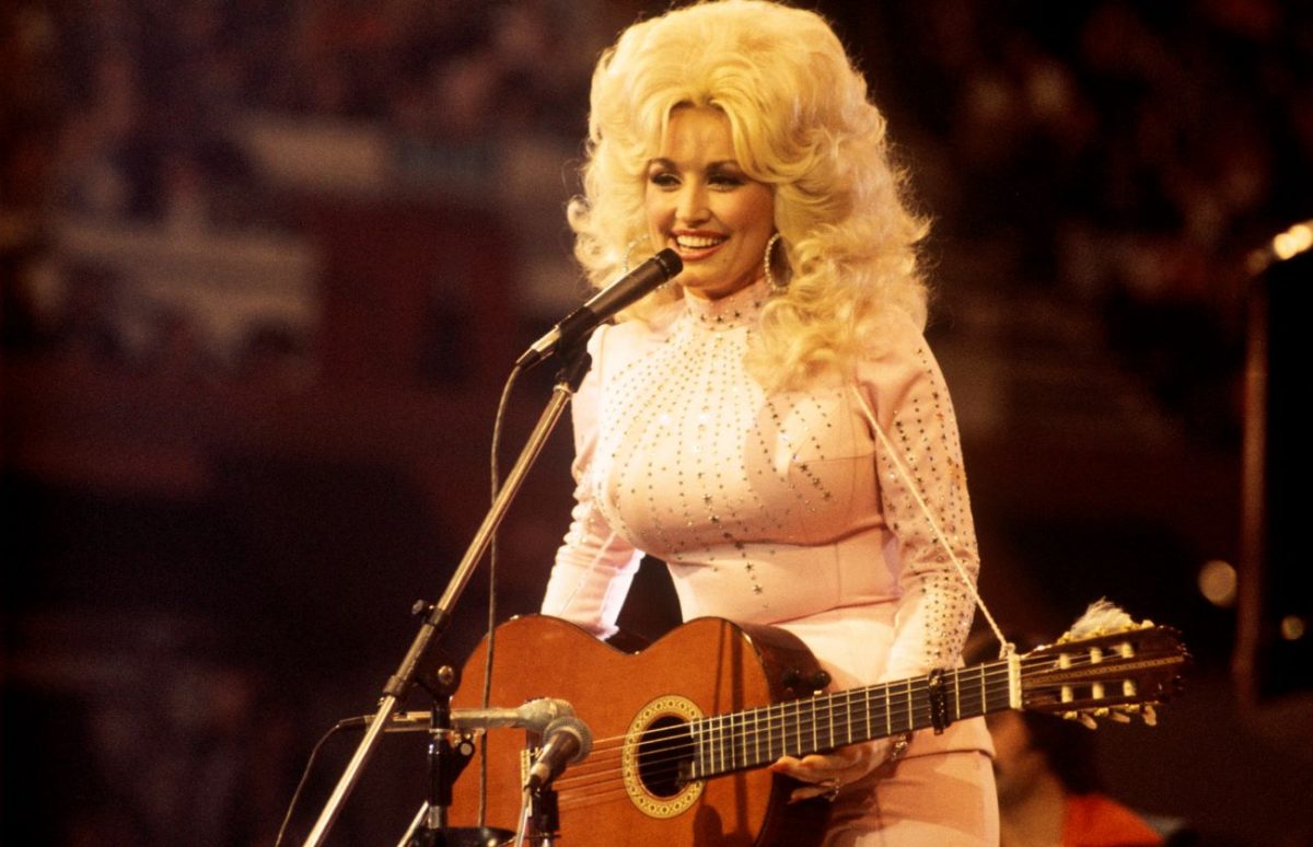 Dolly Parton se tient devant un microphone dans une chemise blanche et une grande perruque de cheveux blonds.