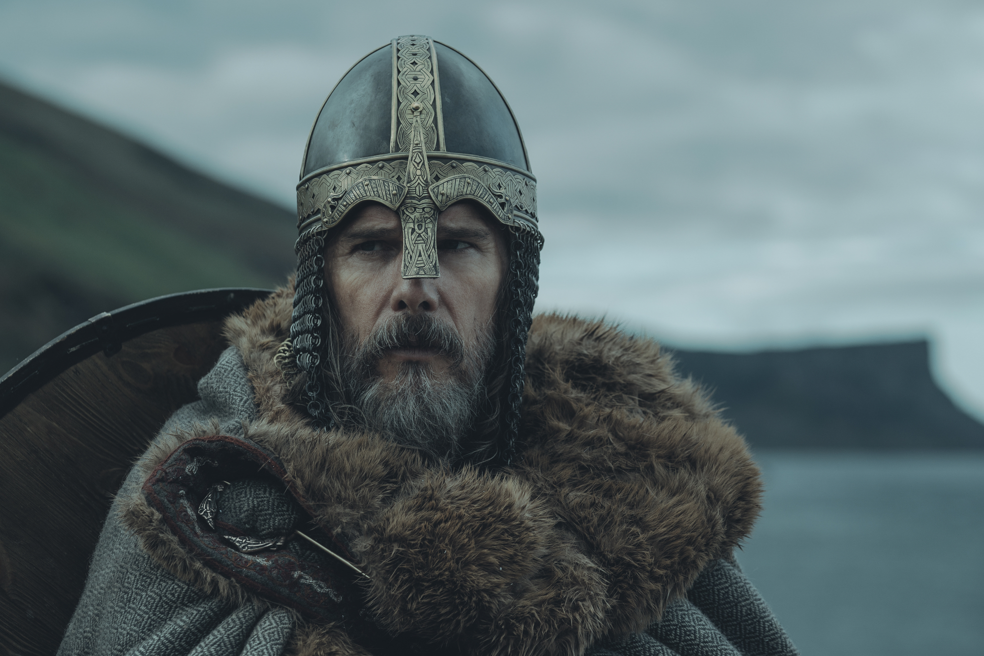 'The Northman' Ethan Hawke dans le rôle du roi Aurvandil War-Raven portant un casque et un manteau devant un paysage de montagne et d'océan