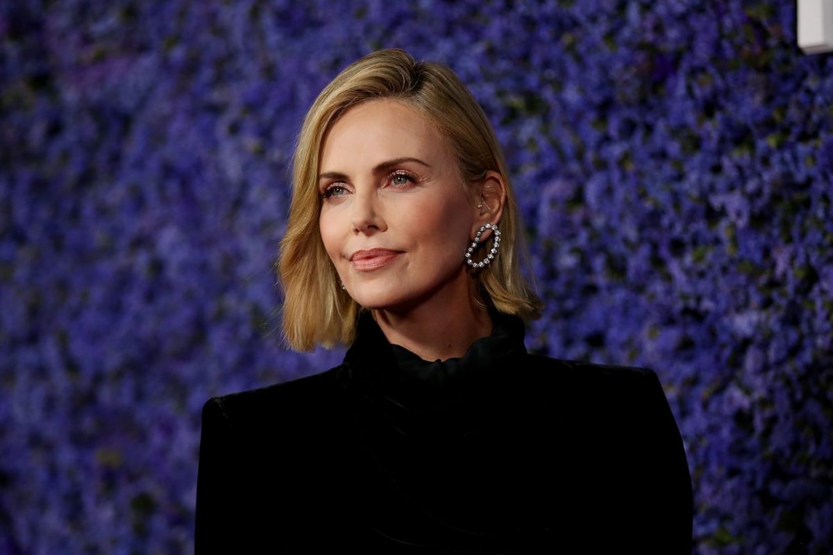 Charlize Theron porte du noir et se tient sur un fond bleu foncé.