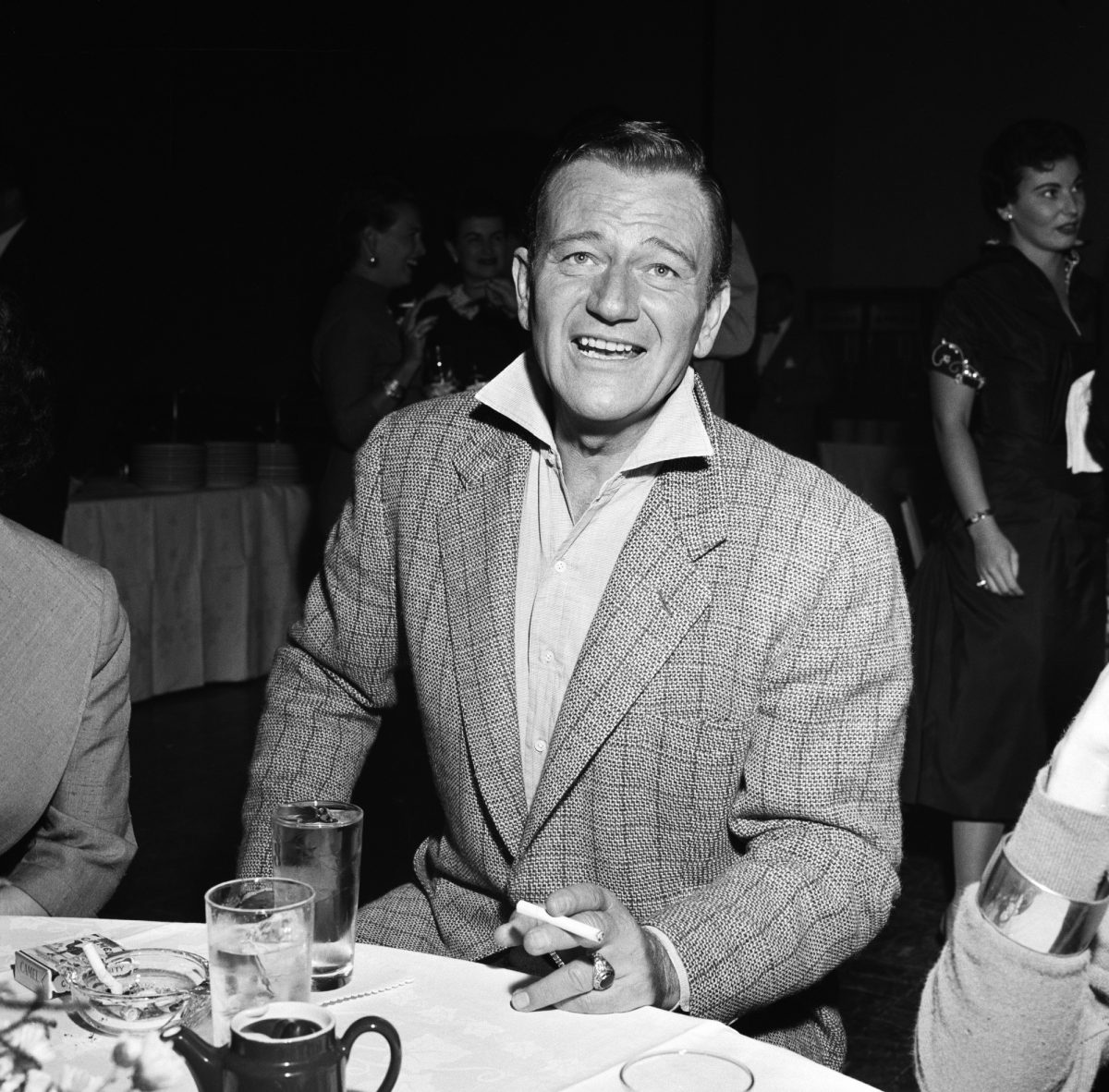 John Wayne, l'acteur qui a donné des conseils à Michael Caine, vêtu d'un costume avec une cigarette à la main, assis à une table