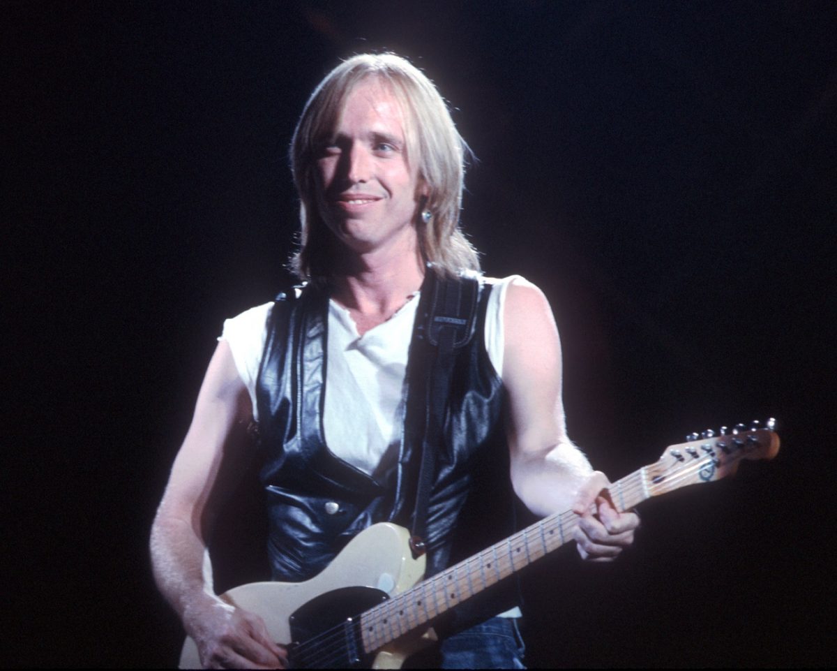 Tom Petty porte un débardeur blanc et un gilet noir et joue de la guitare.