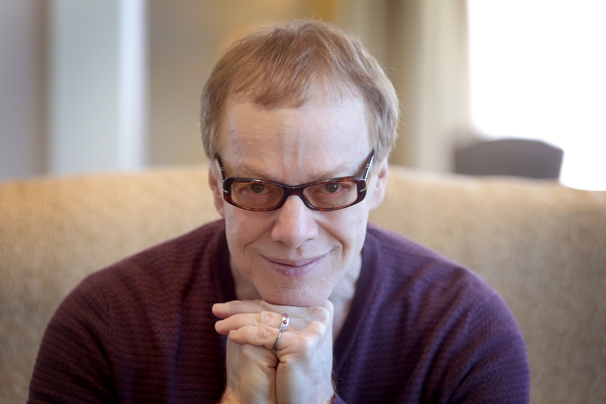 Danny Elfman assis avec un sourire narquois tout en portant des lunettes.