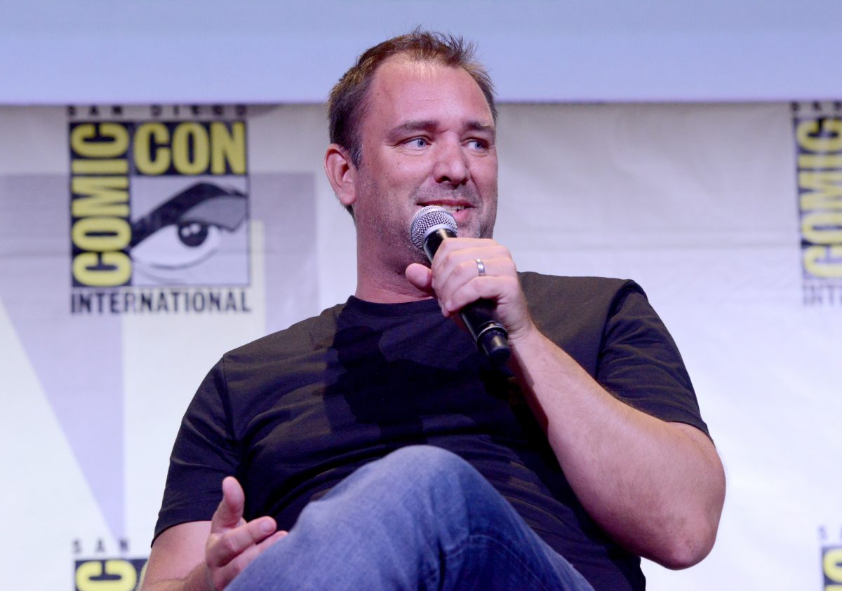 'Cannibale! Le réalisateur du film The Musical Trey Parker portant une chemise à col rond et un jean bleu tenant un microphone devant le logo Comic Con