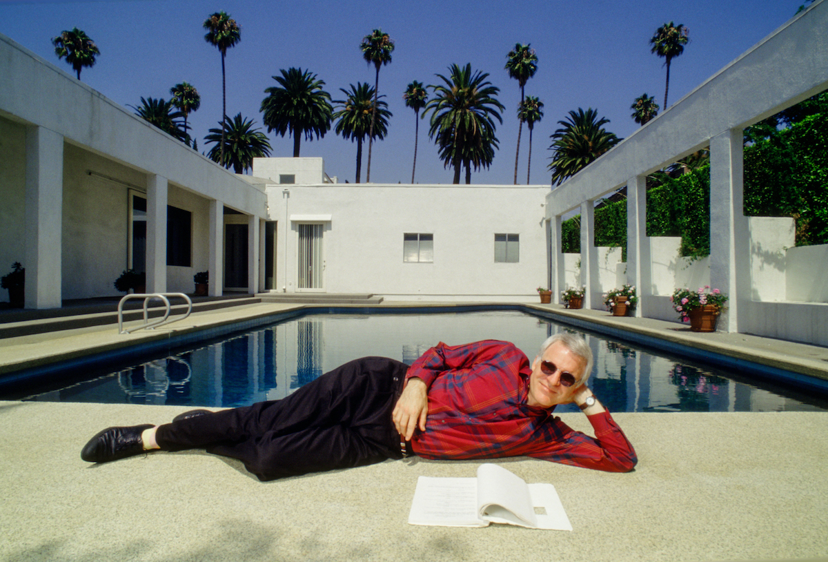 Steve Martin porte une chemise rouge et un pantalon sombre alors qu'il est allongé près d'une piscine