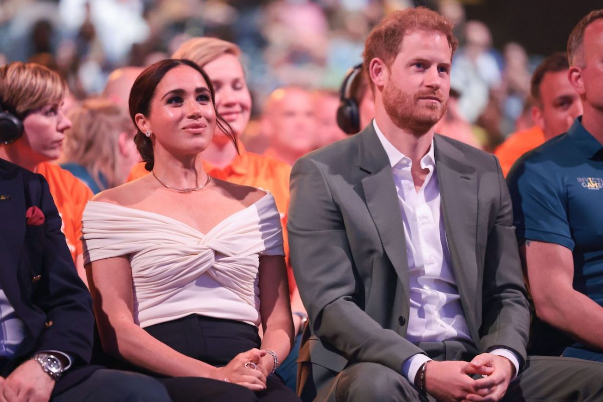 Meghan Markle portant un haut blanc et assise à côté du prince Harry, qui porte un costume gris