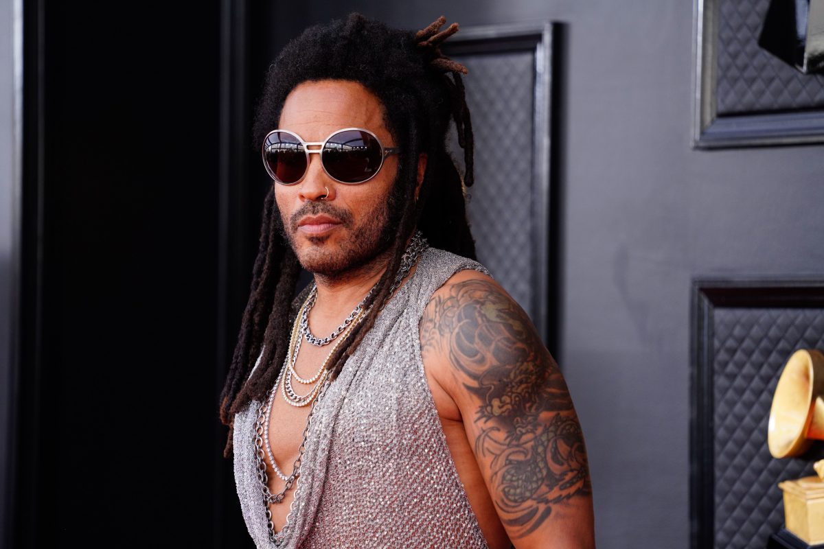 Lenny Kravitz pose sur le tapis rouge tout en portant une chemise argentée transparente, des lunettes de soleil et un pantalon en cuir.