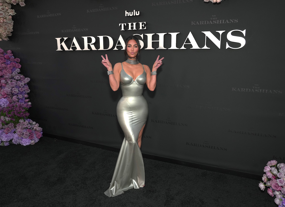 Kim Kardashian pose en faisant une grimace et en lançant deux signes de paix devant un mur qui dit "Hulu The Kardashians".