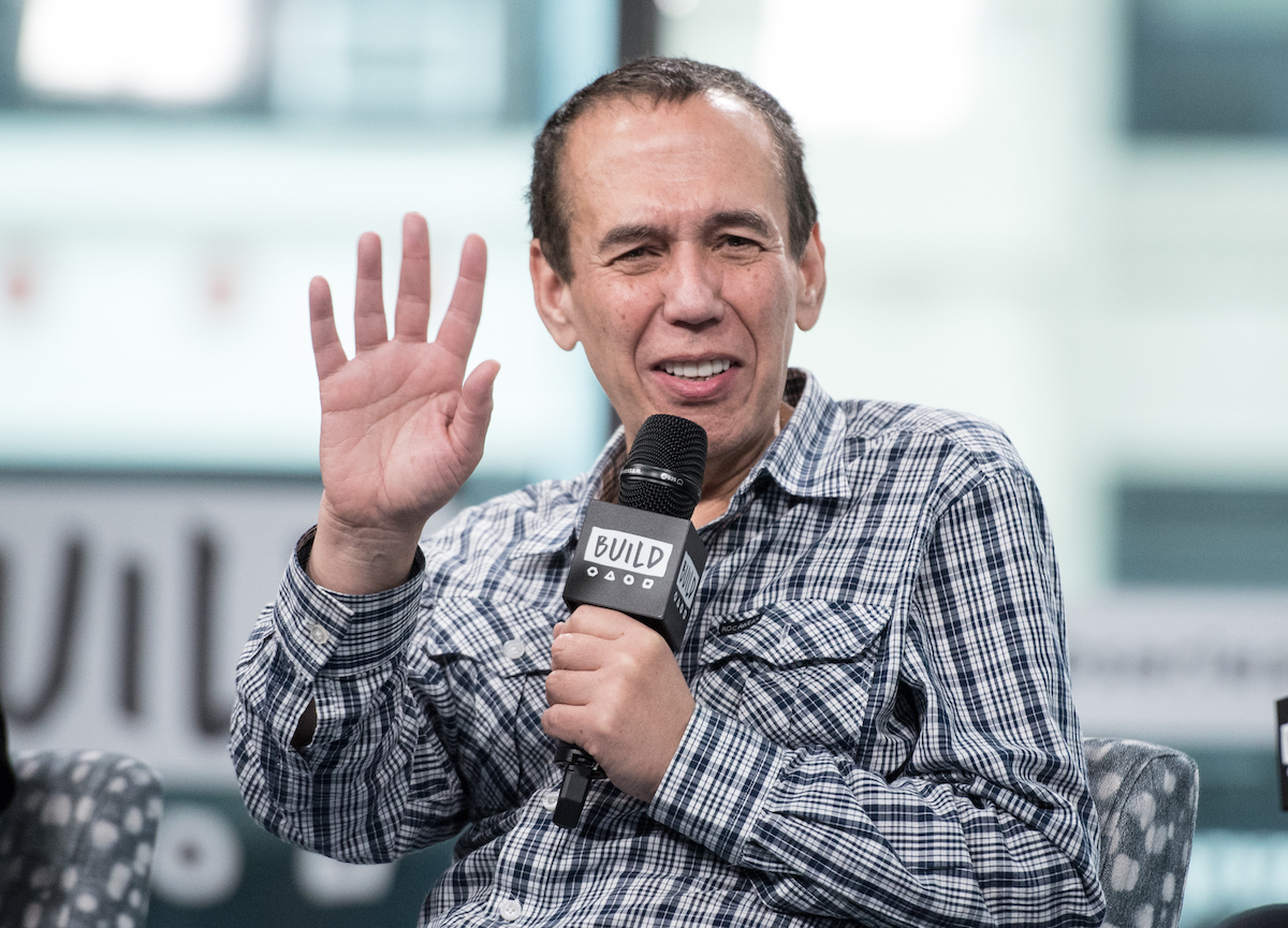 Gilbert Gottfried salue la caméra et tient un microphone.
