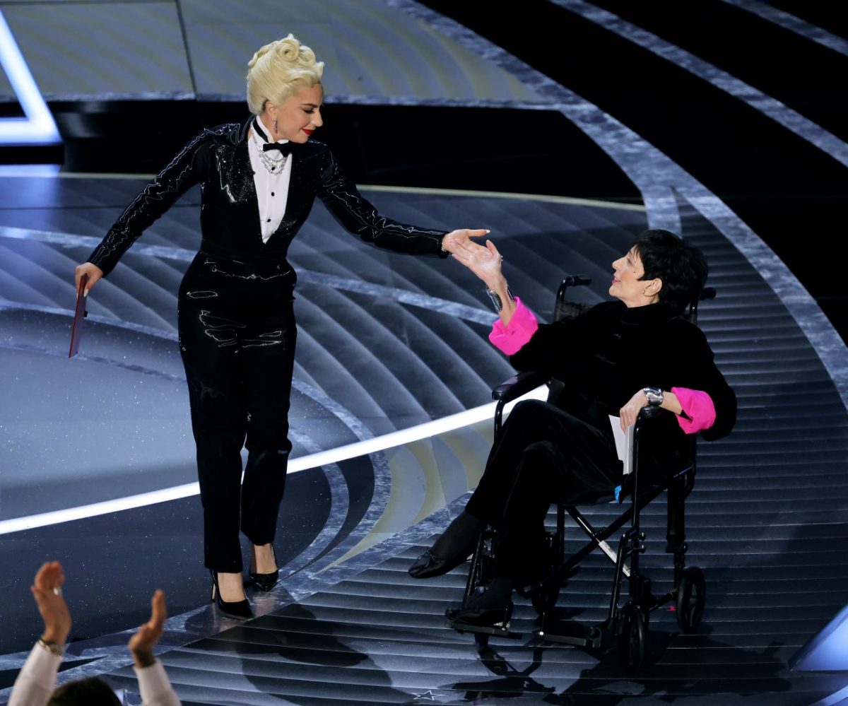Lady Gaga porte un smoking étincelant et touche la main de Liza Minnelli sur la scène des Oscars.  Elle porte une tenue noire avec des manches roses.