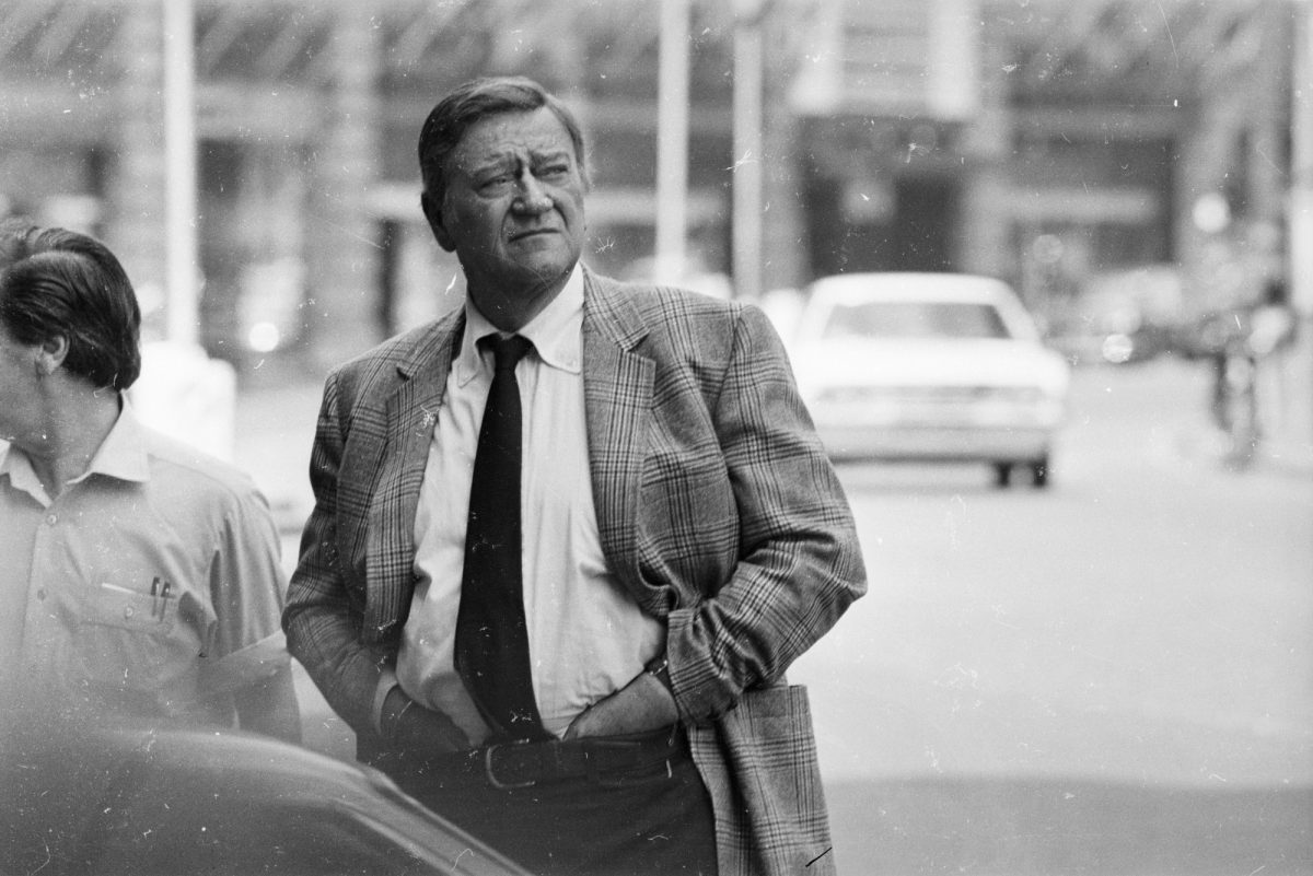John Wayne debout avec sa taille de plus de six pieds portant un costume et une cravate avec une voiture qui s'éloigne en arrière-plan