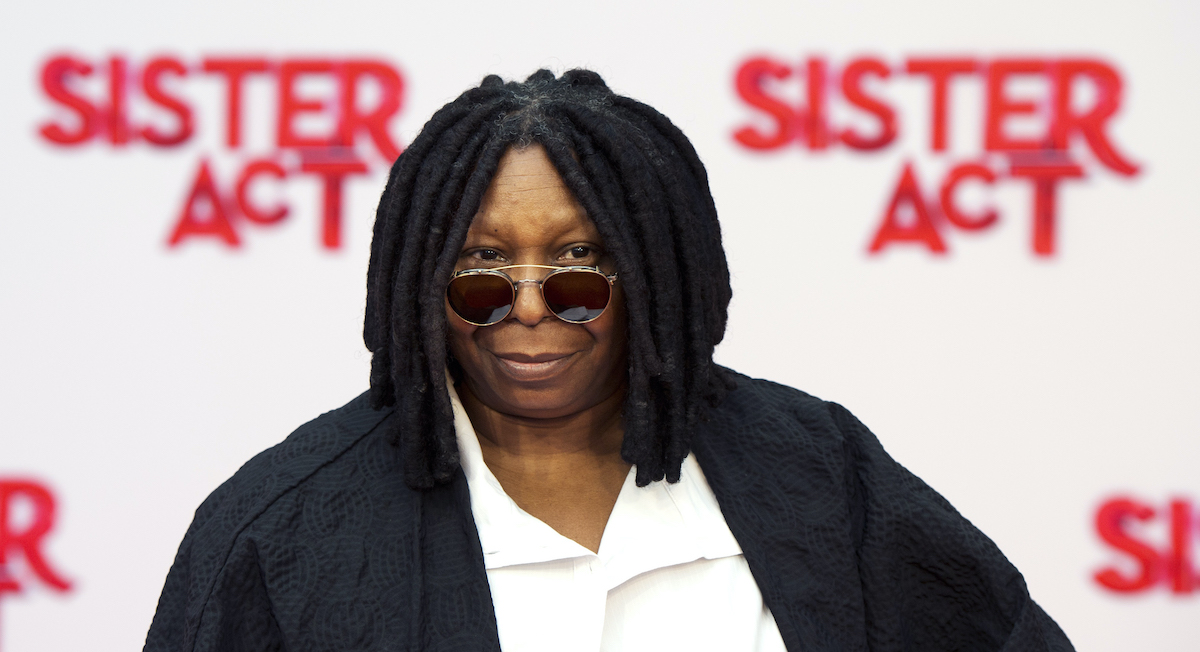 Whoopi Goldberg porte des lunettes de soleil alors qu'elle pose devant le logo 'Sister Act'