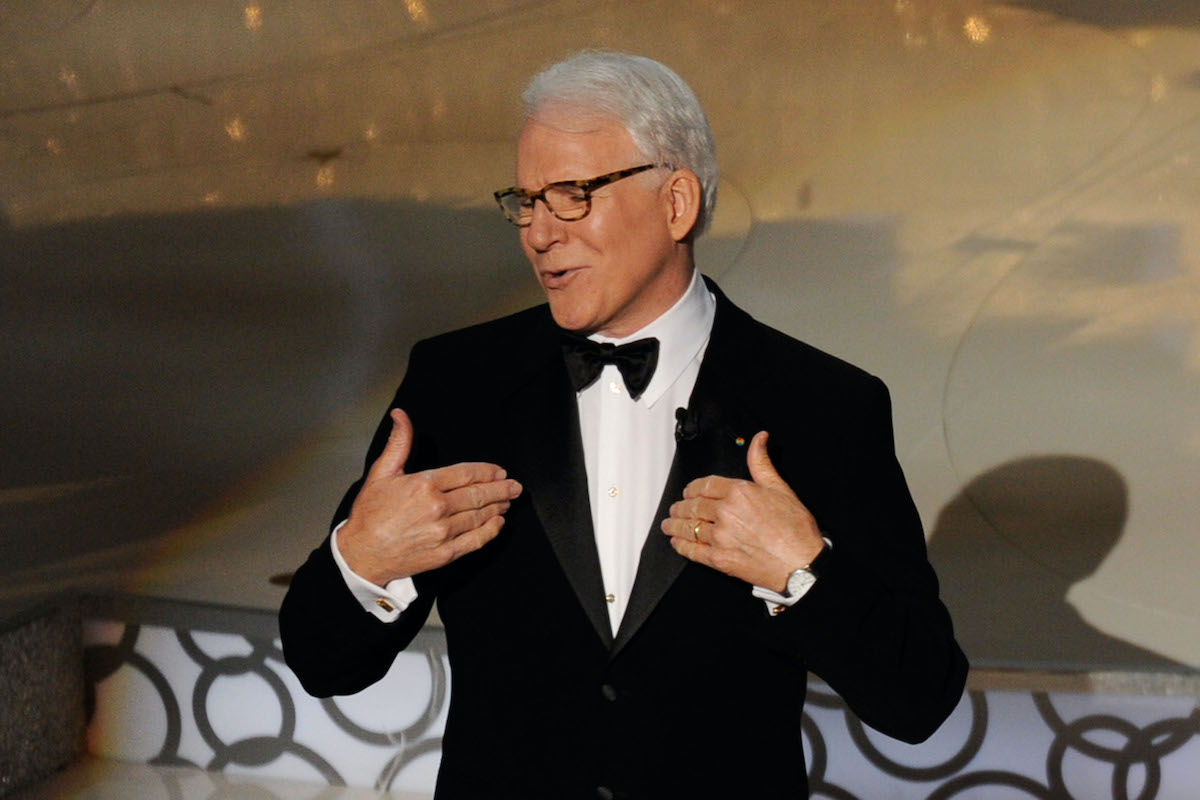 Steve Martin porte un costume et fait un geste vers lui-même sur scène