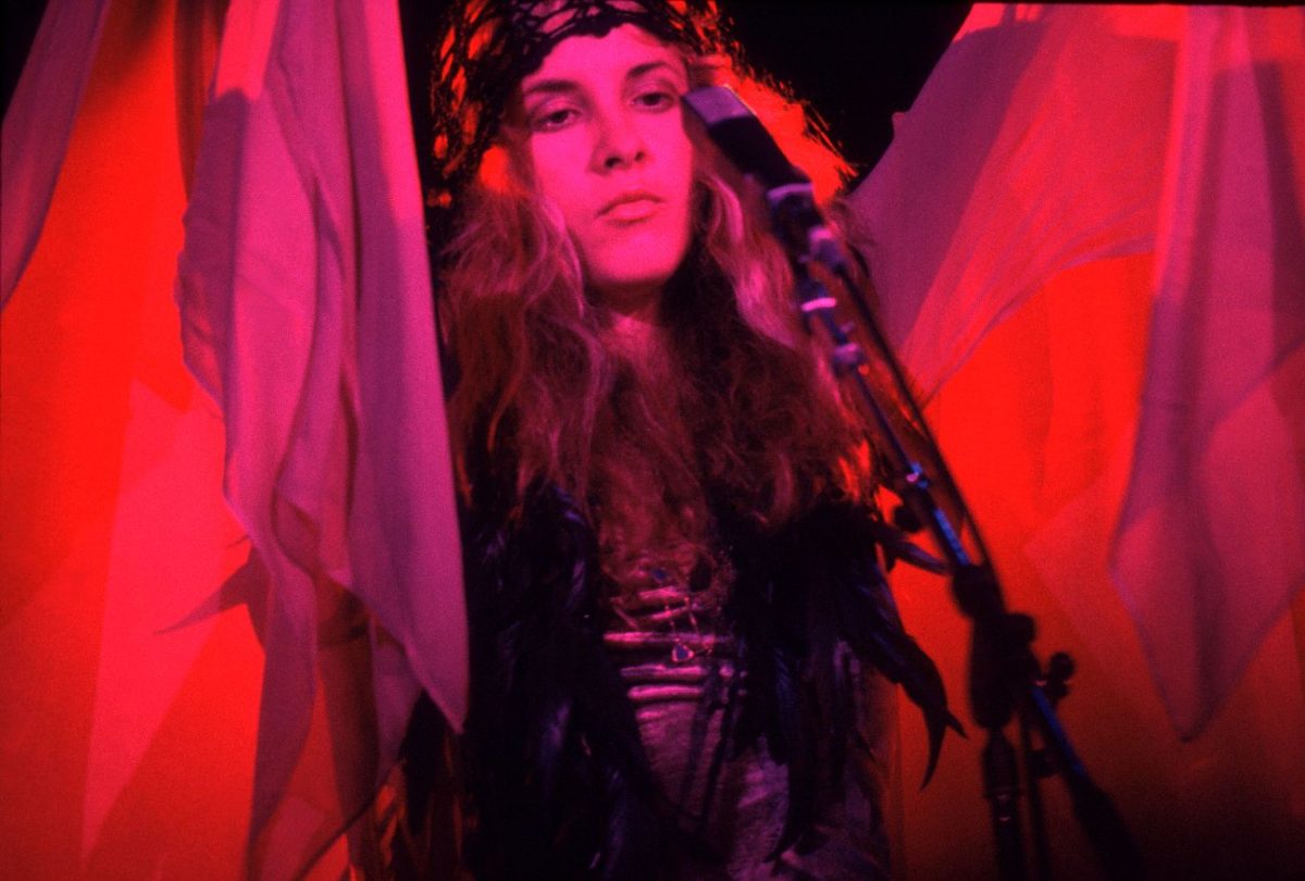 Stevie Nicks soulève son châle au-dessus de sa tête et se tient devant un microphone.