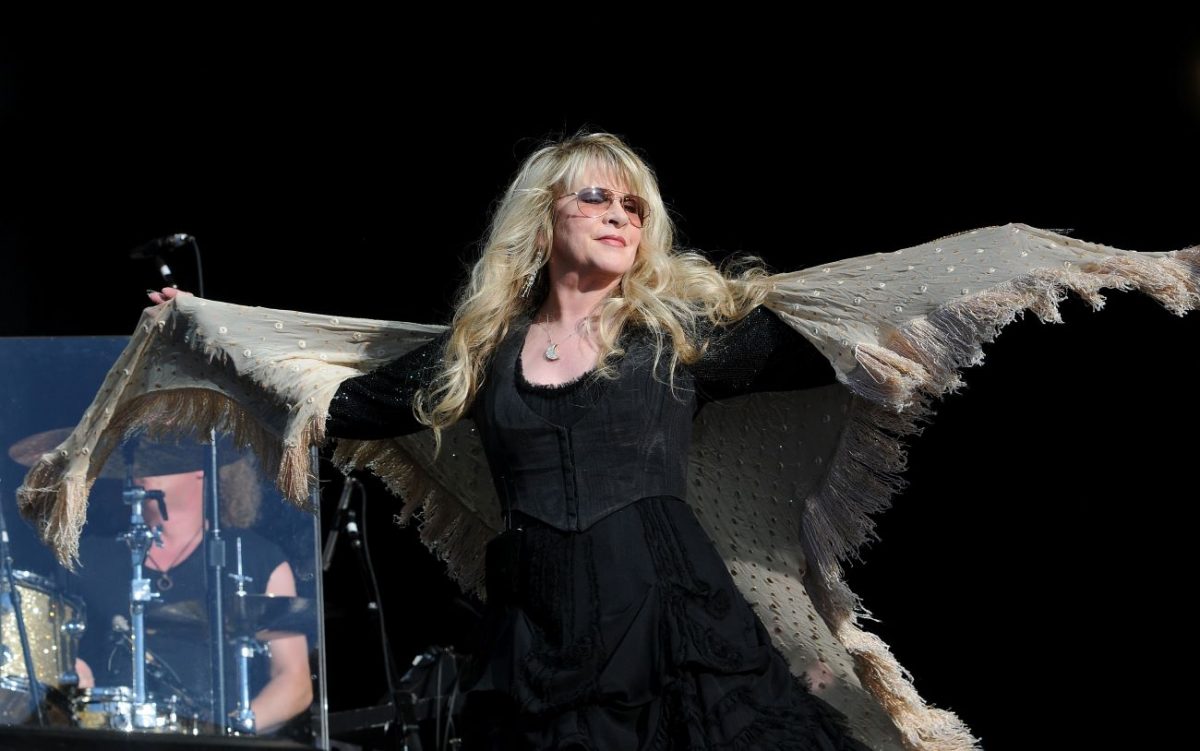Stevie Nicks porte des lunettes de soleil, une robe noire et un châle.  Elle écarte les bras.