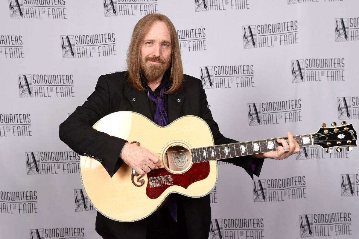 Tom Petty porte un costume noir et tient une guitare. Il se tient devant le fond du Songwriter's Hall of Fame.