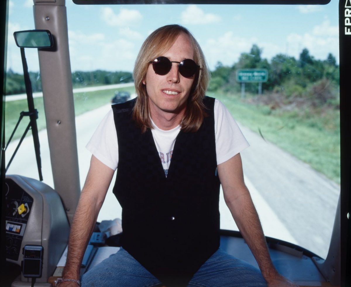 Tom Petty est assis près de la fenêtre avant à l'intérieur d'un bus et porte un gilet noir et des lunettes de soleil