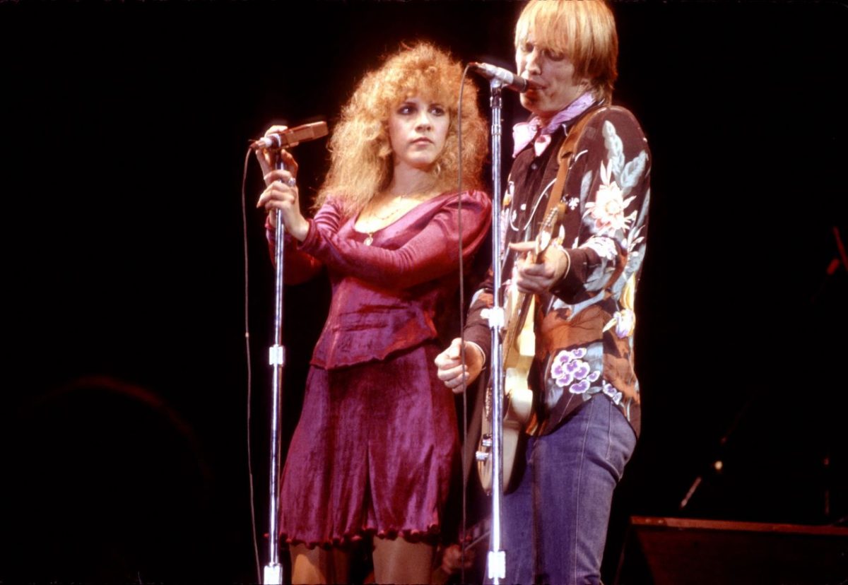 Stevie Nicks porte une chemise et une jupe violettes et tient un microphone.  Tom Petty porte une chemise à fleurs et joue de la guitare en chantant.
