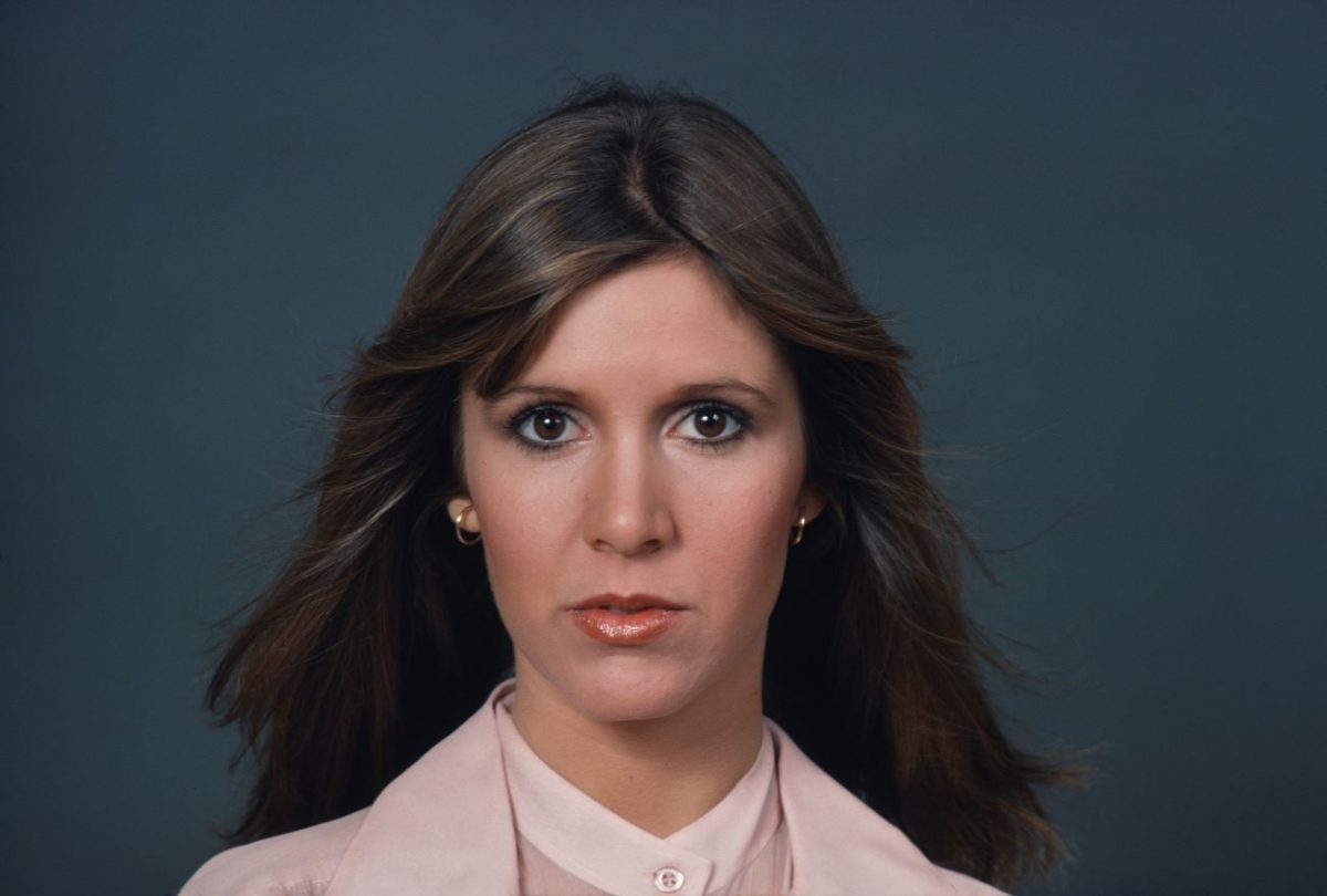 Carrie Fisher porte une tenue rose et se tient devant un fond gris.